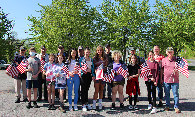 A group of nine middle school students, six men all wearing caps, and a woman stand together on pavement, all holding American flags. There are trees in the background.