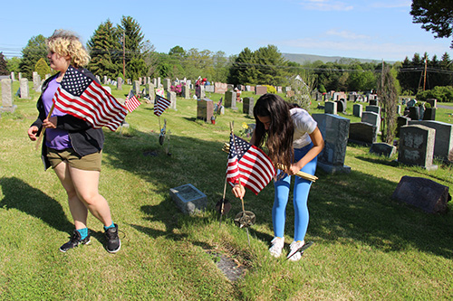 Two young women walk through a cemetery. Both are holding American flags. One, with long dark hair, is putting on on a grave.