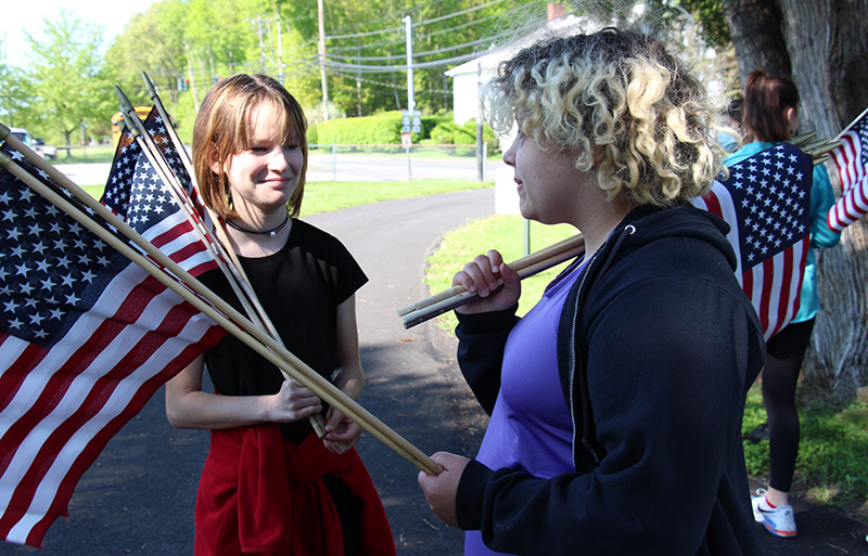 Two middle school girls stand together talking. They are holding flags  on their shoulders.