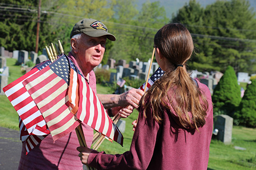 A man in a red shirt holds flats and hands one to a young woman with a long brown ponytail. Her back is to the camera.