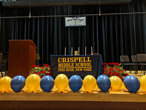 A picture of a stage. In front are blue and gold balloons. There is a table that has a table cloth that says Crispell Middle School. It has five candles on it. Behind it all is a dark blue curtain.