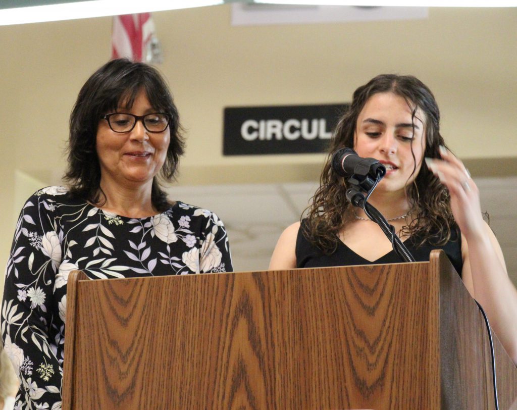 A young woman on the rightt, with long dark hair wearing a black shirt, speaks into a microphone. Next to her is a woman with short dark hair, glasses and a blue and white dress. She is smiing.