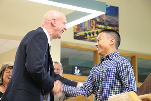 A young man on the right is wearing a blue checked button down shirt. He has short dark hair and is shaking hands with an older man who is wearing a blue jacket. They are both smiling.