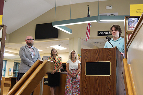 A young man stands at the podium and speaks into a microphone. There is a man on the left, with glasses and a beard, wearing a suit jacket, watching and listening. Two women are in the background listening.