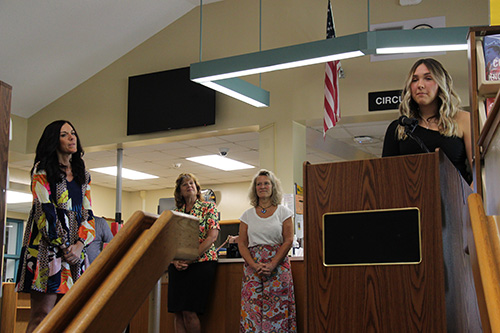 A young woman stands at a podium and speaks into a microphone. There is a woman off to the left wearing a blue printed dress. She has long dark hair and is watching and listening. There are two women in the background listening.