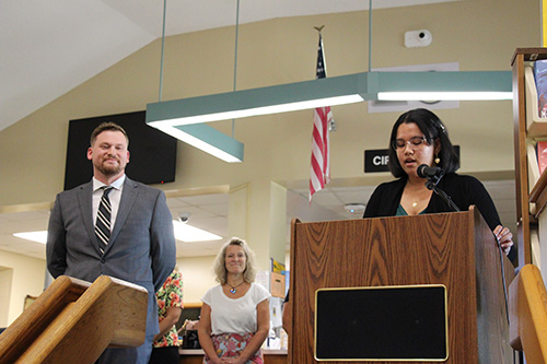 A young woman stands at a podium speaking into a microphone. She has long dark hair and is wearig a black sweater. There is a man to her left, wearing a jacket and tie. He is smiling. A woman is in the background listening.