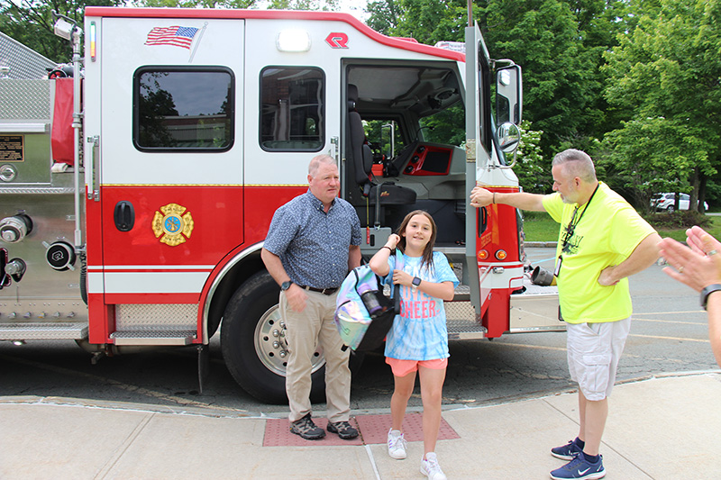 A fifth-grade girl wearing a light blue shirt and pink shorts puts her backpack on as she exits a fire engine. There is a man standing to greet her wearing a yellow shirt, and a man in the background wearing a blue shirt and tan pants.