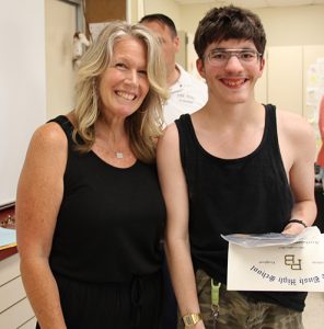 A woman with shoulder-length blonde hair dressed in a black shirt and pants has her arm around a high school boy also dressed in black. He is holding a certificate and is smiling.