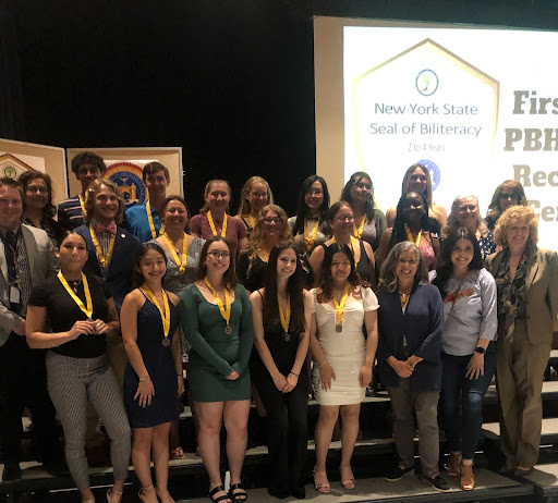 A group of 18 high school students and several teachers stand in three rows. The students all have gold ribbons around their necks with medallions on them.