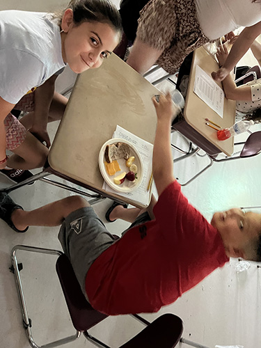 A group of three students sit at a table working on a project. On the table is a plate filled with food - bread, cheese, banana and apple slices.