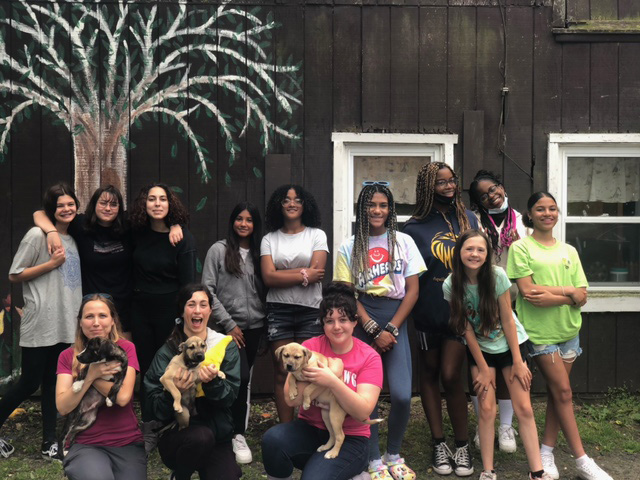 A dark building is the background with a tree drawn on it. In front is a group of 13 people, middle school students in the back and three in front holding puppies. All are smiling.