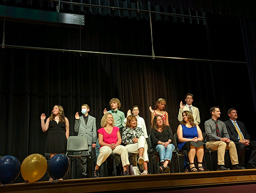 Front row has adults sitting . Behind them are six middle school studets standing, holding their right hand up taking a pledge.