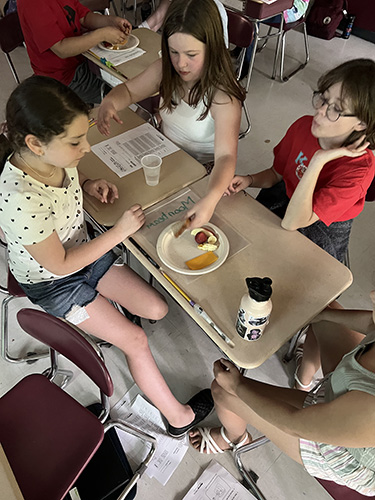 A group of three elementary students work at desks together. On the desk is a plate with food on it, including cheese, bread and apple.