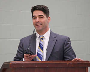 A man with dark hair smiles. He is at a podium holding a microphone. He is wearing a gray suit, white shirt, and blue striped tie.