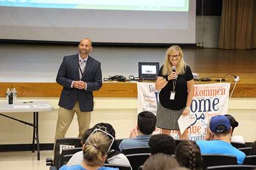 A man in a blue jacket, dress shirt and khaki pants is on the left, and a woman in black shirt, black and white skirt with long blonde hair and glasses stand in front of an auditorium filled with people speaking to them.