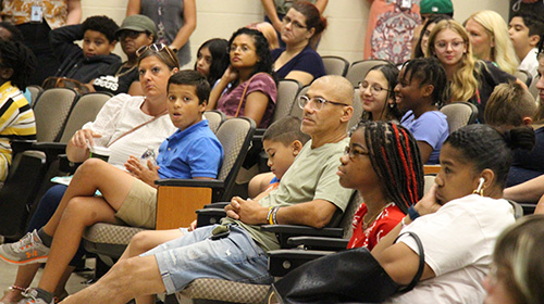 A group of parents and middle-school age children sit in an auditorium listening to a speaker.