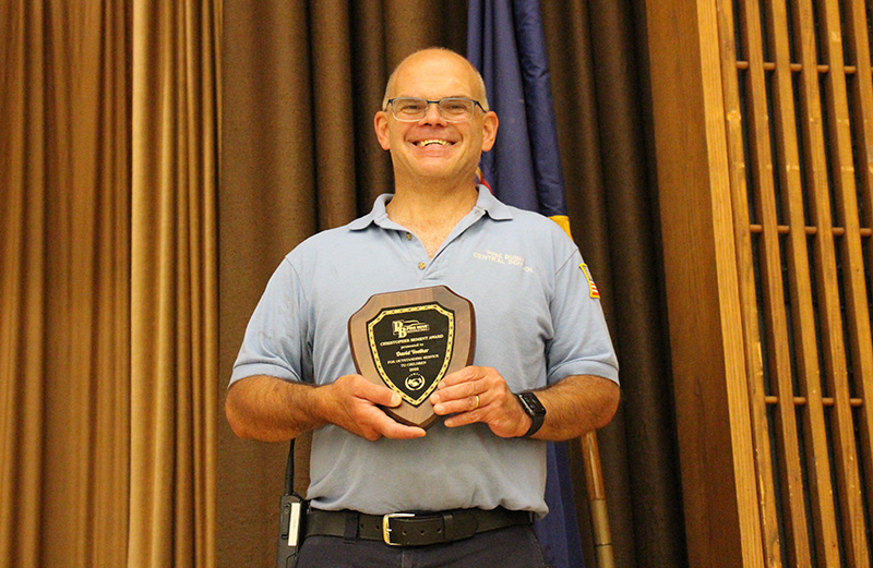 A man with a big smile, wearing glasses, alight blue short-sleeve shirt, stands on a stage holding a plaque