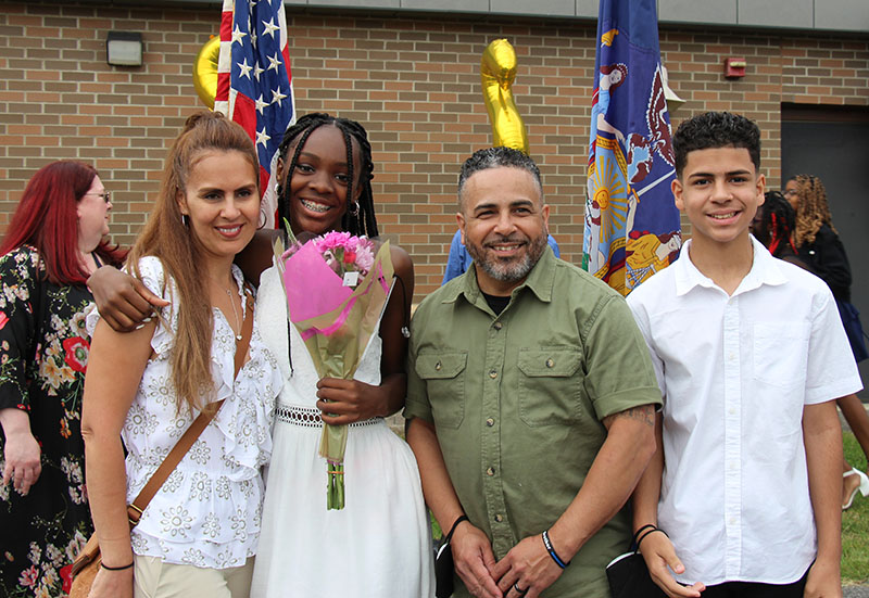 A Young woman, second from left, wearing a white dress, with long black braids, holds pink flowers. On the left is a woman with long lighter hair, wearing a white shirt. On the other side is a man with short dark hair, a beard and wearing a green short-sleeve shirt. On the right is a young man wearing a white shirt. He is smiling and has short dark hair.