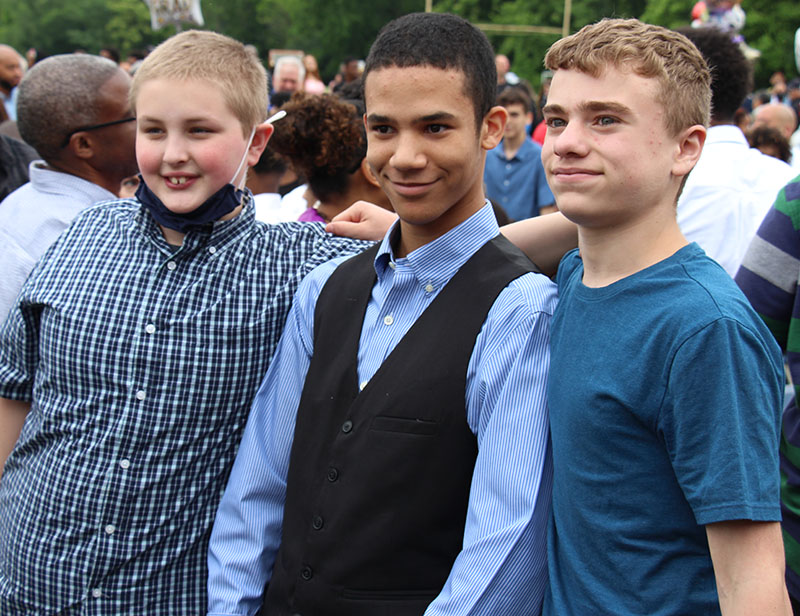 Three eighth grade boys stand together smiling. The kid on the left has short light hair, is wearing a blue plaid shirt and is smiling. The boy in the center is wearing a long-sleeved blue button-down shirt and dark vest. He has dark short hair and is smiling. The boy on the right is wearing a blue tshirt and has short blonde hair. He is smiling too. They are outside.