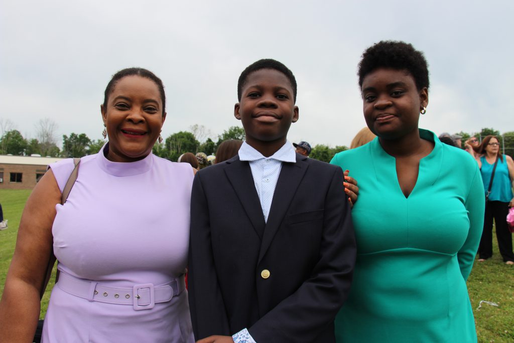 Two women, one on the left wearing a light purple dress and th eothe ron the right wearing a green dress, stand on either side of a young man wearing a white dress shirt and dark jacket. They are outside and smiling.