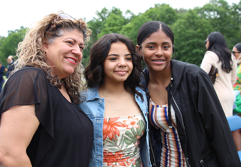 A young woman stands in the center of two other women. She is wearing a flowered dress and blue jacket. She has long dark hair. The woman on the left is wearing a black shirt and has shoulder-length light hair. On the other side is another young woman wearing a printed shirt and black jacket. She has long dark hair that is pulled back. They are all smiling.