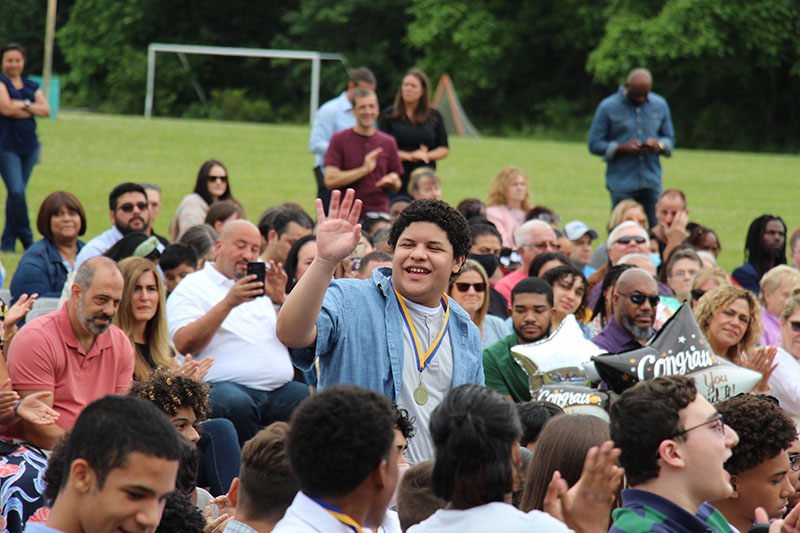 A young man wearing a blue shirt and a medal around his neck, stands up in a crowd of kids and waves. He has short dark hair and is smiling.