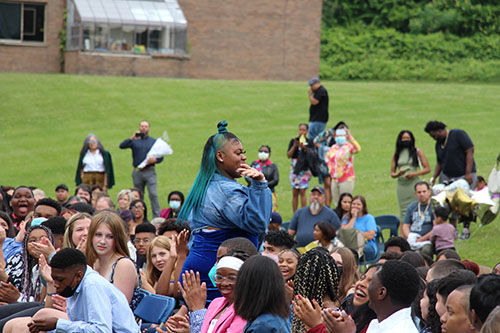 A young woman wearing a blue jacket stands up among a group of kids. she waves. She has long dark hair with green streaks in it.