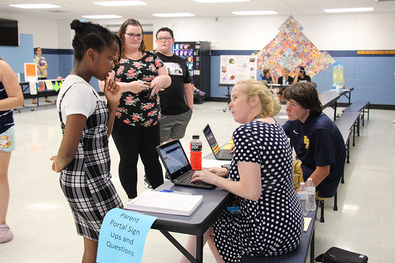 A woman in a blue and white polkadot dress sits at a table using a laptop. She is looking up at a middle school student, who has her dark hair pulled back. She is wearing a plaid dress and white shirt. There are others standing by the table.