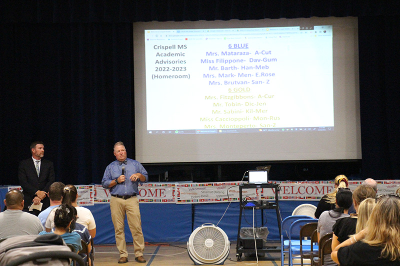 Two men stand to the left of the photo, with a large screen next to them. There are people in chairs listening to them and watching the presentation.