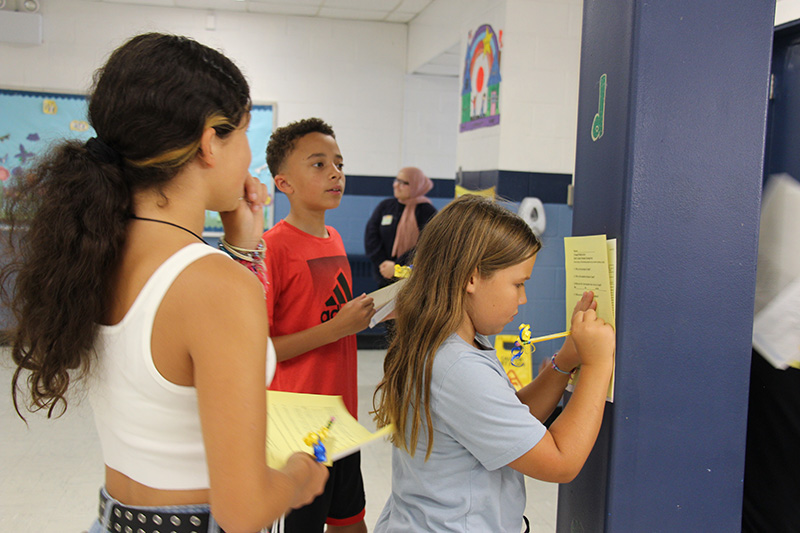 A group of three middle school students all have yellow paper and pencils. One, with a blue short-sleeve shirt and long blonde hair, writes on the paper as she leans it against a blue wall. The other two students stand next to her. One is a boy with a red shirt and very short dark hair, the other is a girl with long dark hair pulled back in a ponytail, wearing a white shirt.