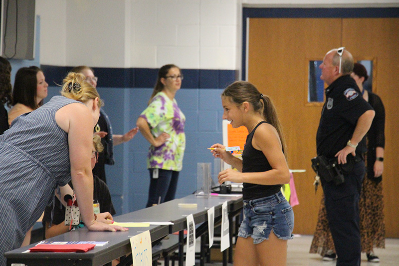 A young middle school girl, with shorts and a tank top, dark hair pulled back into a ponytail, talks to two people at a table. The people at the table are getting volunteers for their Students Making a Difference Club.