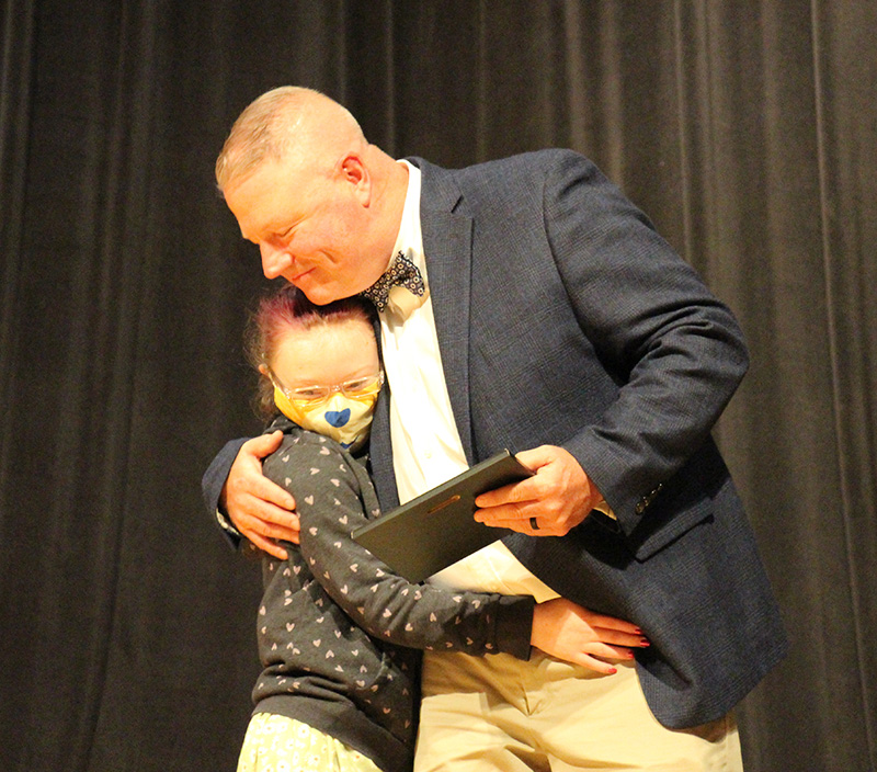 A man wearing tan pants, blue jacket, white shirt and bow tie hugs a little girl who has her long hair pulled back and is wearing a black and white jacket and a mask with an animal's nose on it.