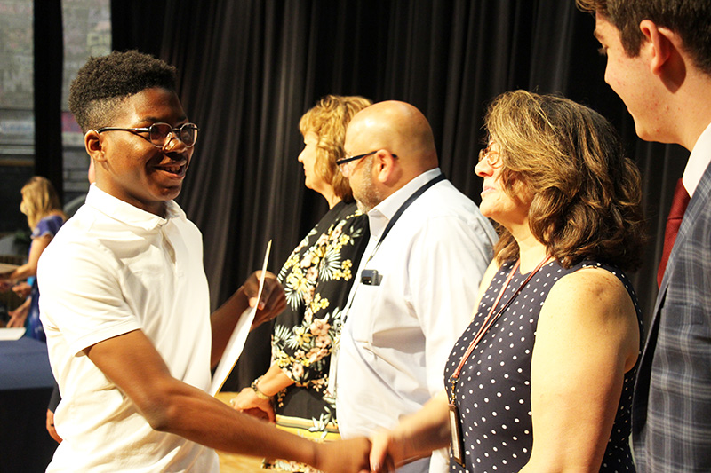 A young man wearing a white polo shirt shakes hands with a woman in a blue and white polka dot dress. She has shoulder-length dark hair. There are two other adults on the stage next to her.