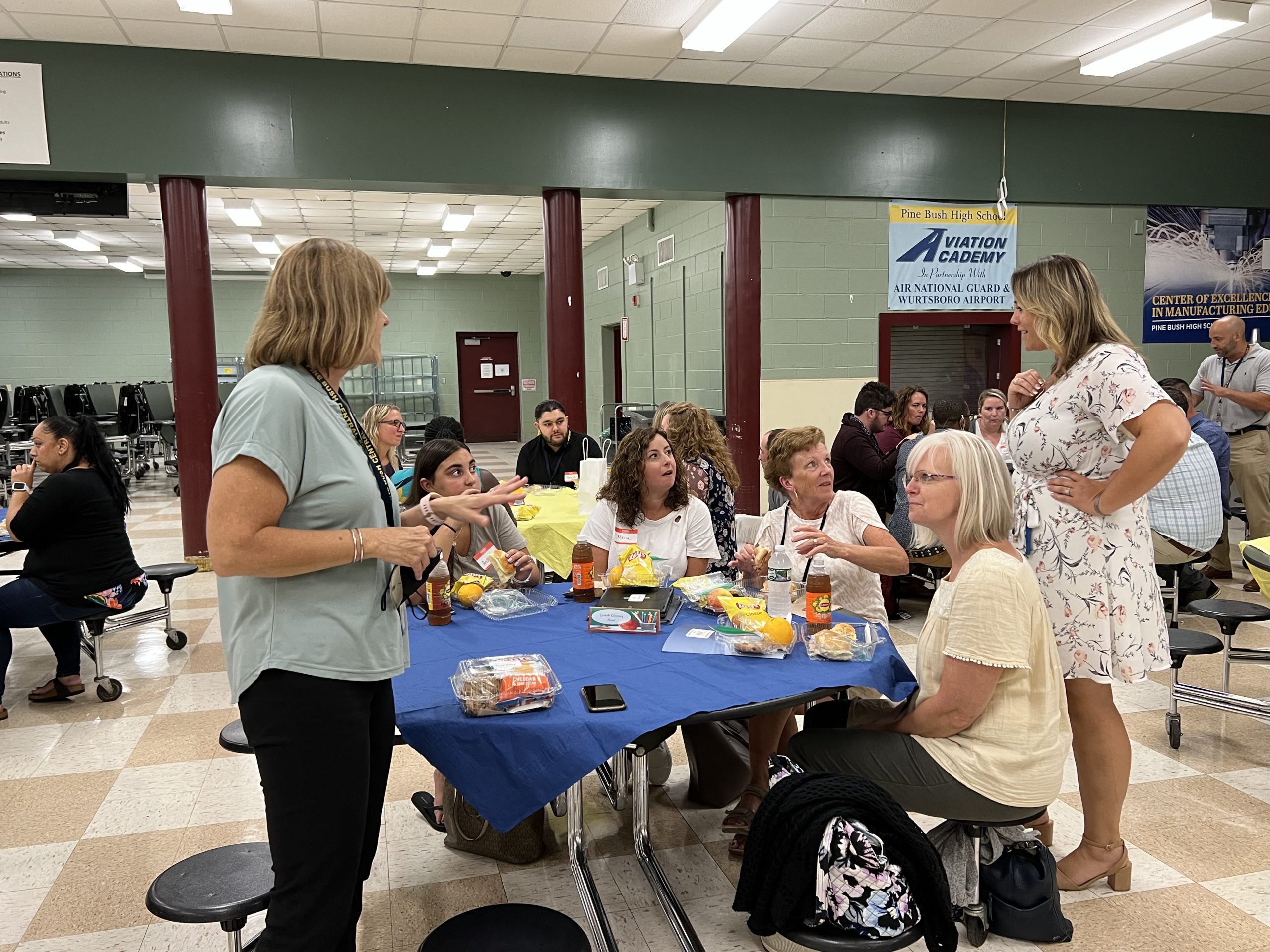 A table with a blue tablecloth on it. There are four women sitting at the table with wraps in front of them. Two women are standing and talking with those witting. There are others around in the cafeteria.