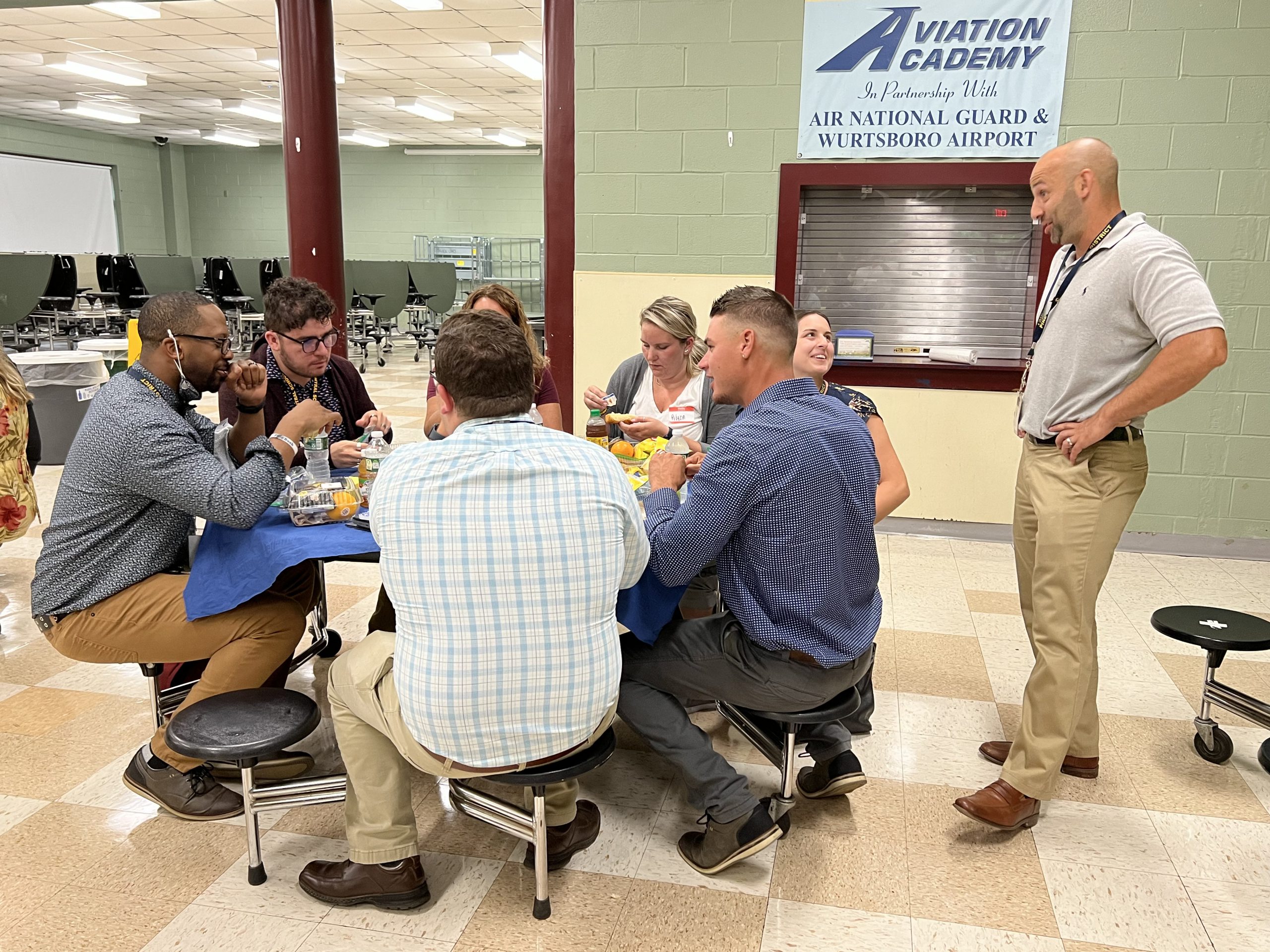 A group of men and women sit at a round table with blue tablecloth eating and talking. One man is standing talking to the group.