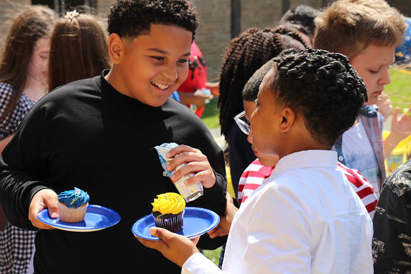 Two fifth-grade boys talk to each other. They are both holding blue plates that contain a cupcake - one blue and one yellow. The boy facing the camera is smiling.