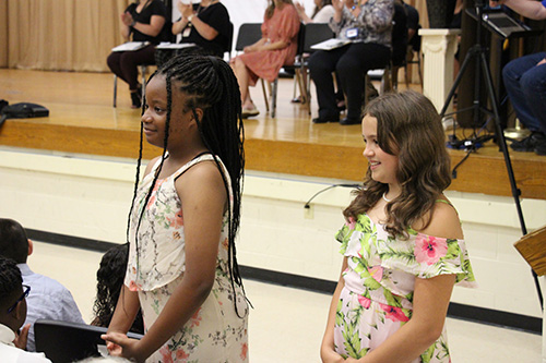 Two fifth-grade girls, both with long dark hair, stand at their seats in an auditorium. 