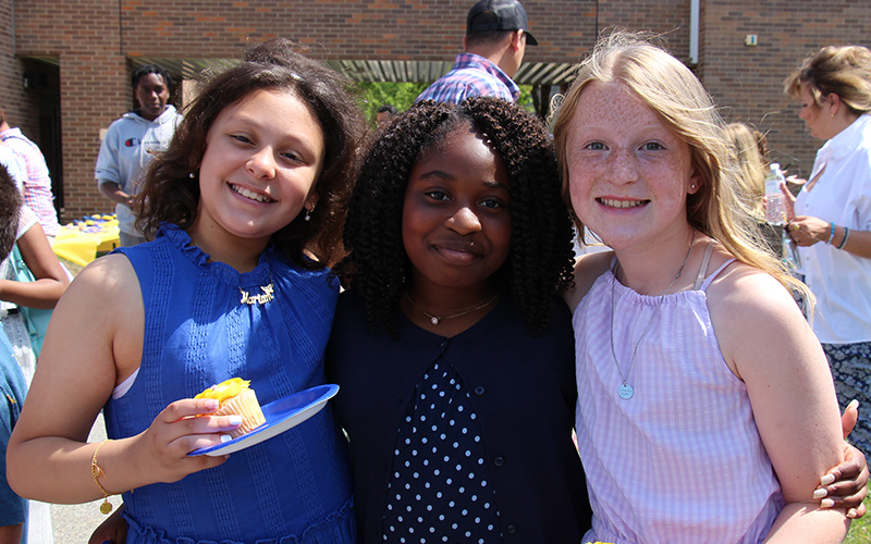 Three fifth-grade girls smile, with their arms around each other. The girl on the left has dark hair and is wearing a royal blue shirt. She is holding a cupcake. The girl in the middle has long dark hair and is wearing a blue an dwhite polka dot dress and dark sweater. The girl on the right has long blonde hair and is wearing a white shirt. They are all smiling.