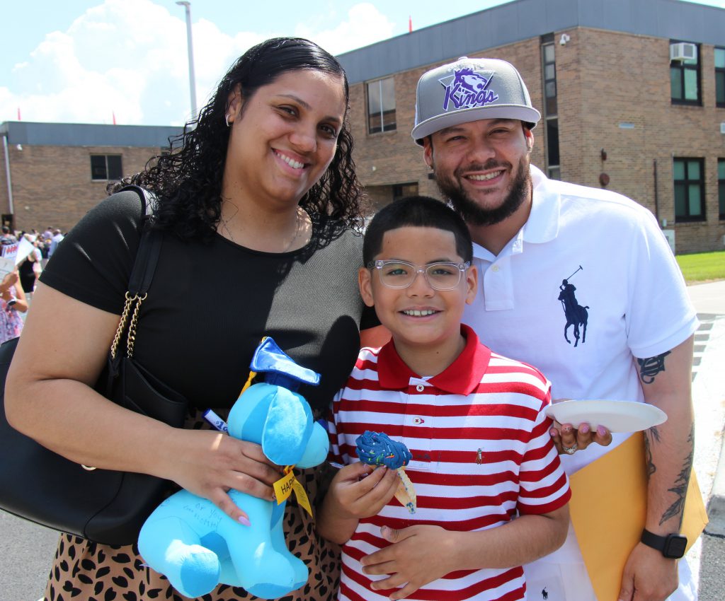 A fifth grade boy with short dark hair is wearing a red and white polo shirt and holds a blue cupcake. He is standing in front of a woman with long dark hair and a man wearing a white shirt and baseball cap. All are smiling.