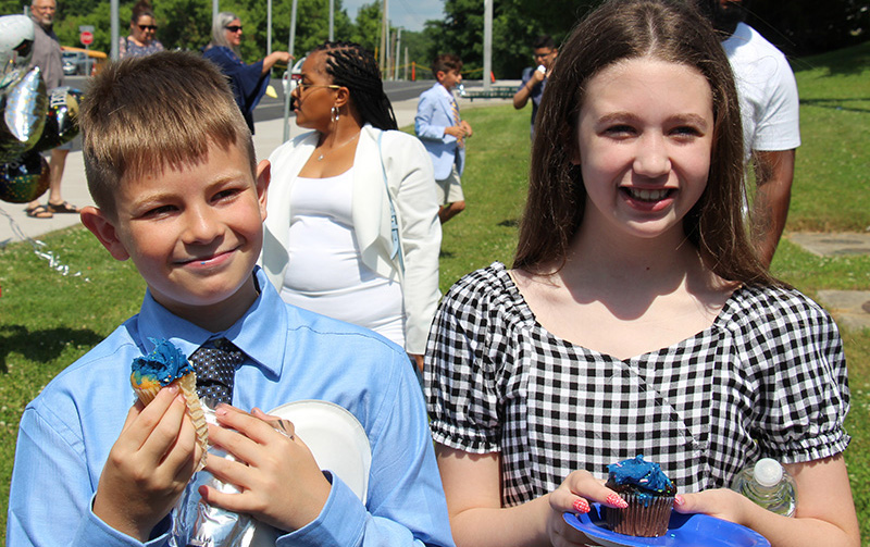 Two fifth-grade students - a boy wearing a blue long-sleeved shirt and blue tie, and a girl with long dark hair wearing a black and white checked dress - smile. Each is holding a cupcake.