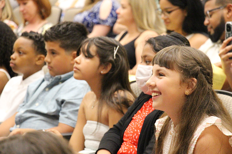 A row of fifth-grade students sit listening to a speaker. The girl on the end has long brown hair and is smiling.