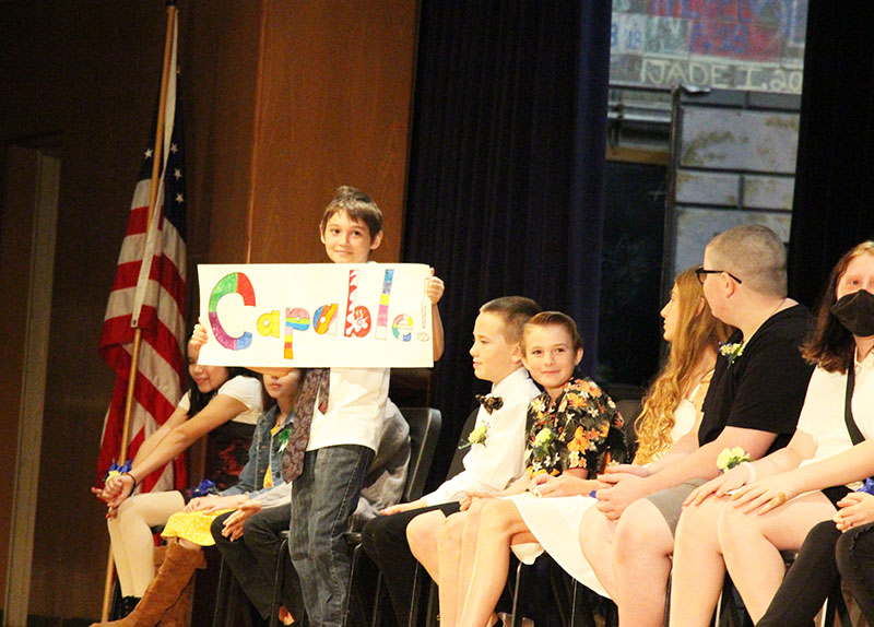 A fifth-grade boy stands up holding a paintd sign that says Capable. His classmates are sitting around him.