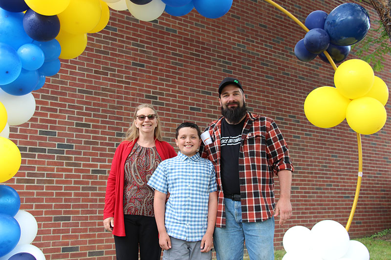 A circle of blue and yellow and white balloons with three people standing in the center. A woman with a red sweater and long blonde hair smiles. In the center is a fifth-grade boy with dark hair, wearing a blue plaid dress shirt, and a man wearing blue jeans, plaid shirt, baseball cap. All are smiling.