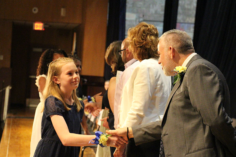 A fifth-grade girl with long blonde hair shakes hands with a man wearing a suit.