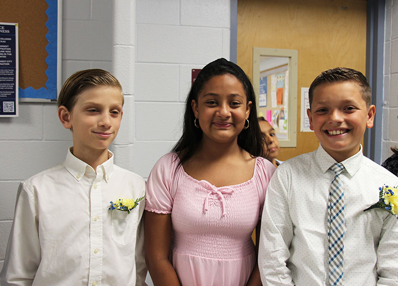 Three fifth-grade students stand together smiling. A boy in a white shirt with a yellow flower pinned on, a girl with dark shoulder-length hair wearing a pink dress, and a boy with a white shirt and patterned tie.