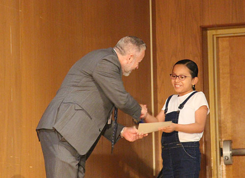 A man in a suit shakes the hand of a fifth-grade girl who is smiling. He is handing her a certificate.