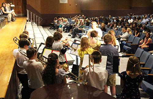 A group of fifth-grade students play instruments. The view is from behind them and there are many people in the audience watching and listening.