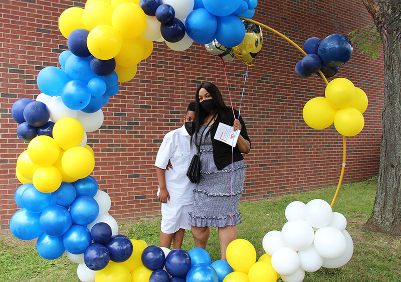 A woman wearing a light blue dress stands with a fifth-grade boy, who is wearing a white shirt and shorts. They are standing  surrounded by a circle of blue, yellow and white balloons.