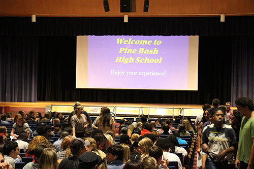 A large screen at the front of an auditorium sayd Welcome to Pine Bush High School. There are many people seated in front of it.