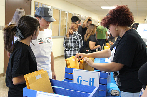 A student and adult stand in front of a  table with boxes labeled with different letters of the alphabet. Another student thumbs through the envelopes in one of the boxes marked G-H.